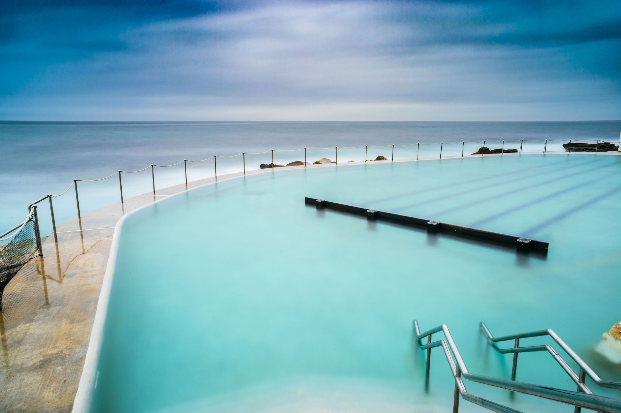 The calm water of Bronte Baths on a clear day, the open sea beyond the pool wall