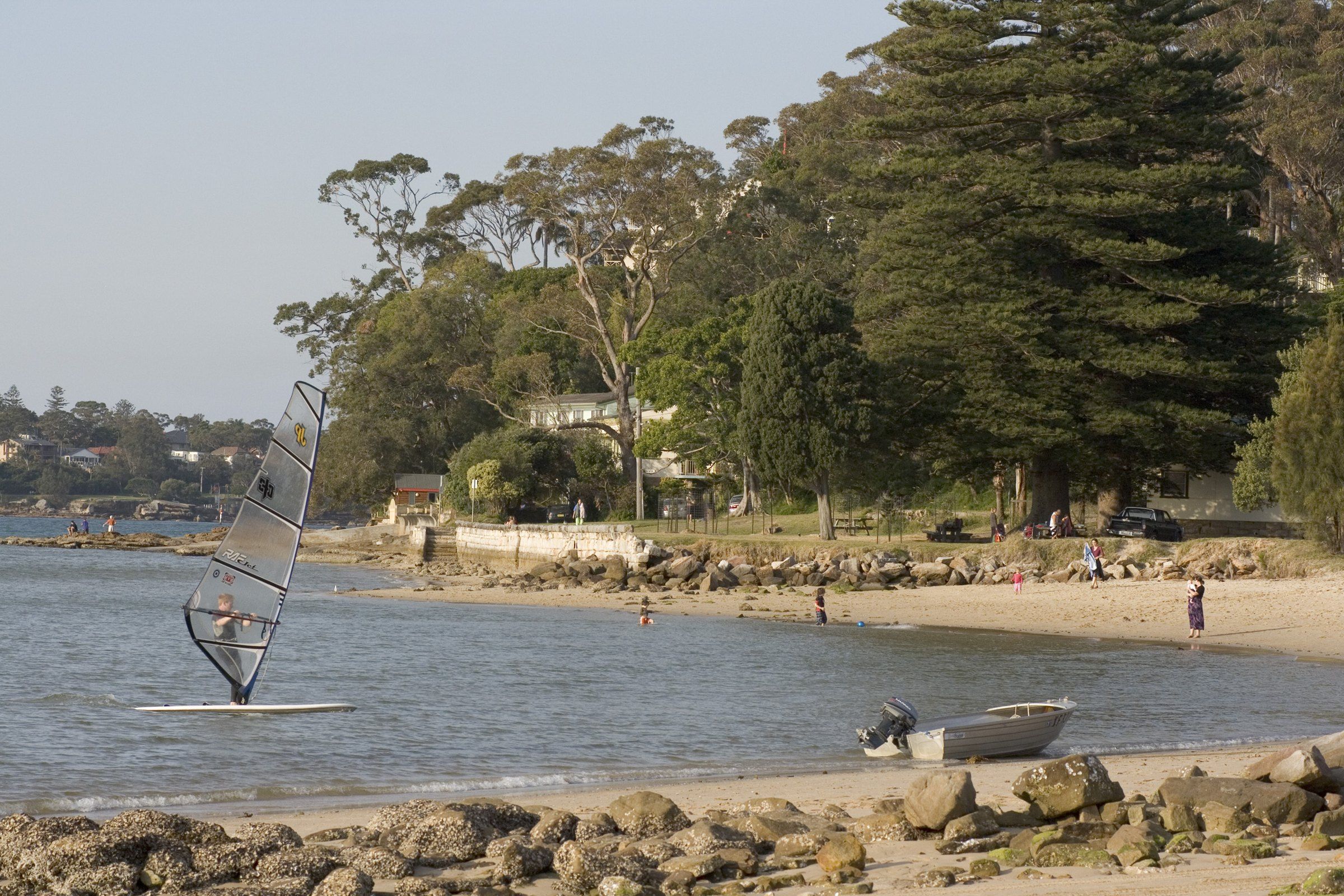 A windsurfer on calm water off a beach at Bundeena, trees behind