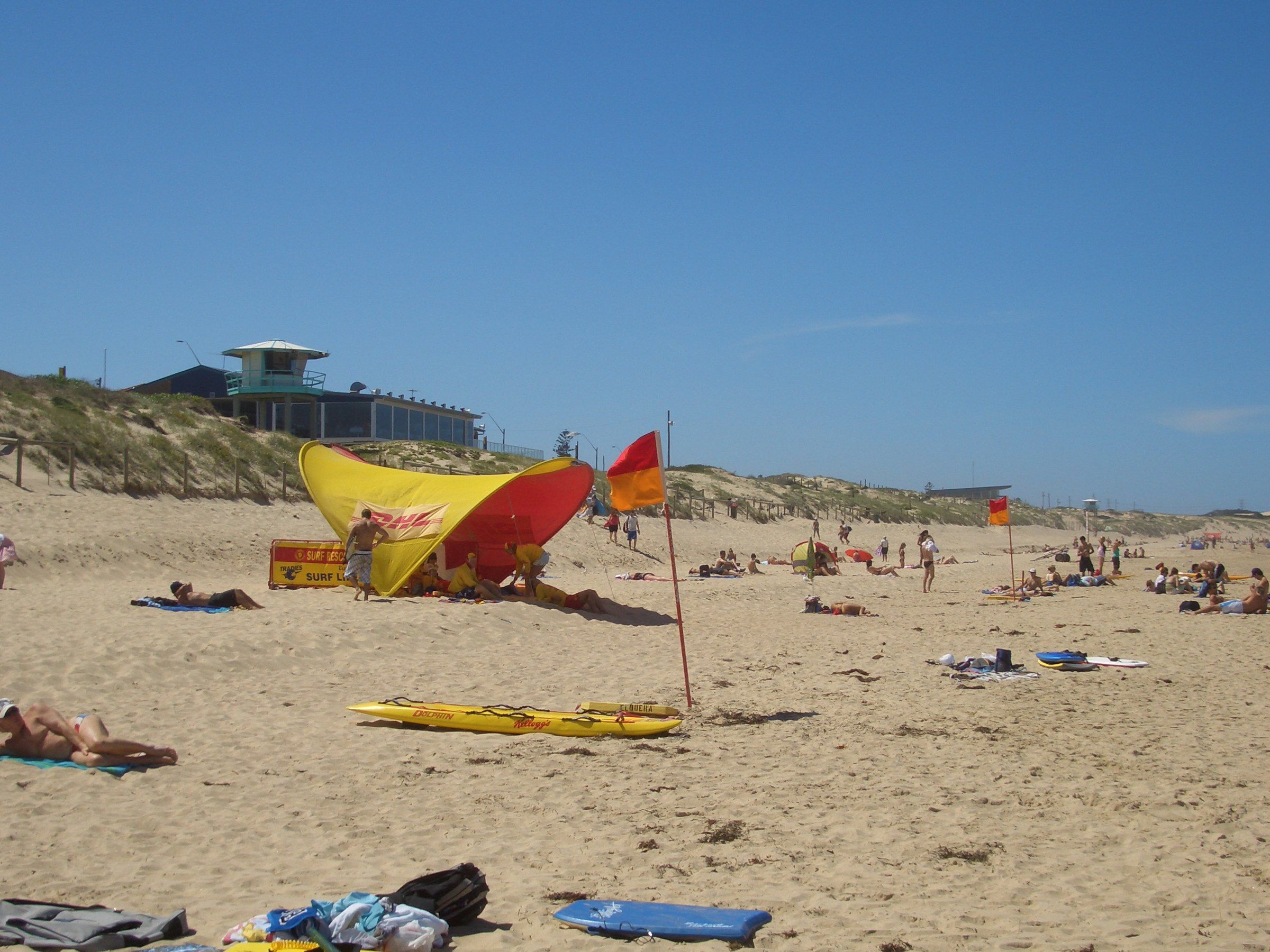 Red and yellow flags and a lifeguard tent on Elouera Beach on a sunny day, the surf club behind the dunes