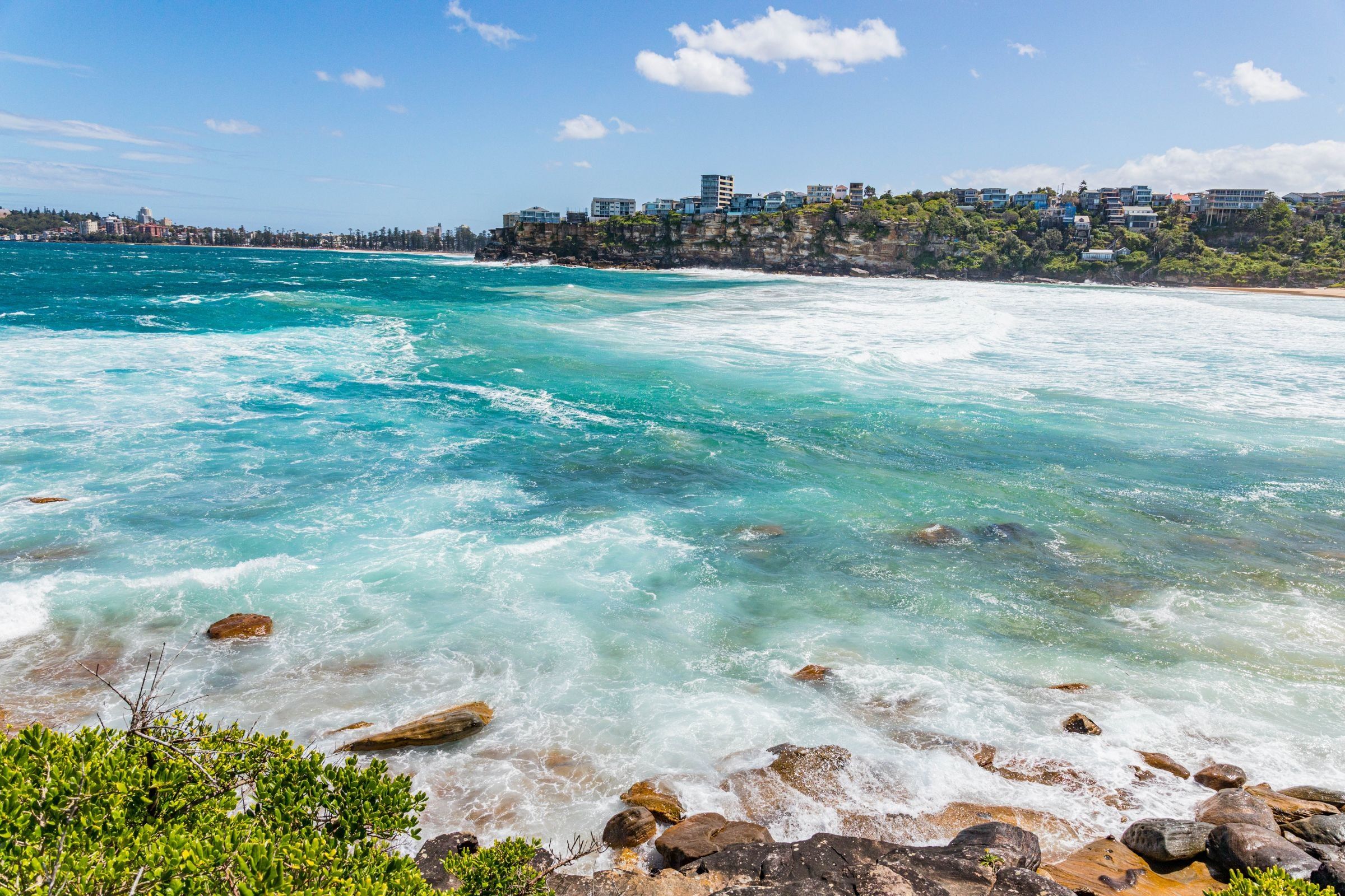 Turquoise surf rolling into Freshwater Beach on a sunny day, the headland and houses behind