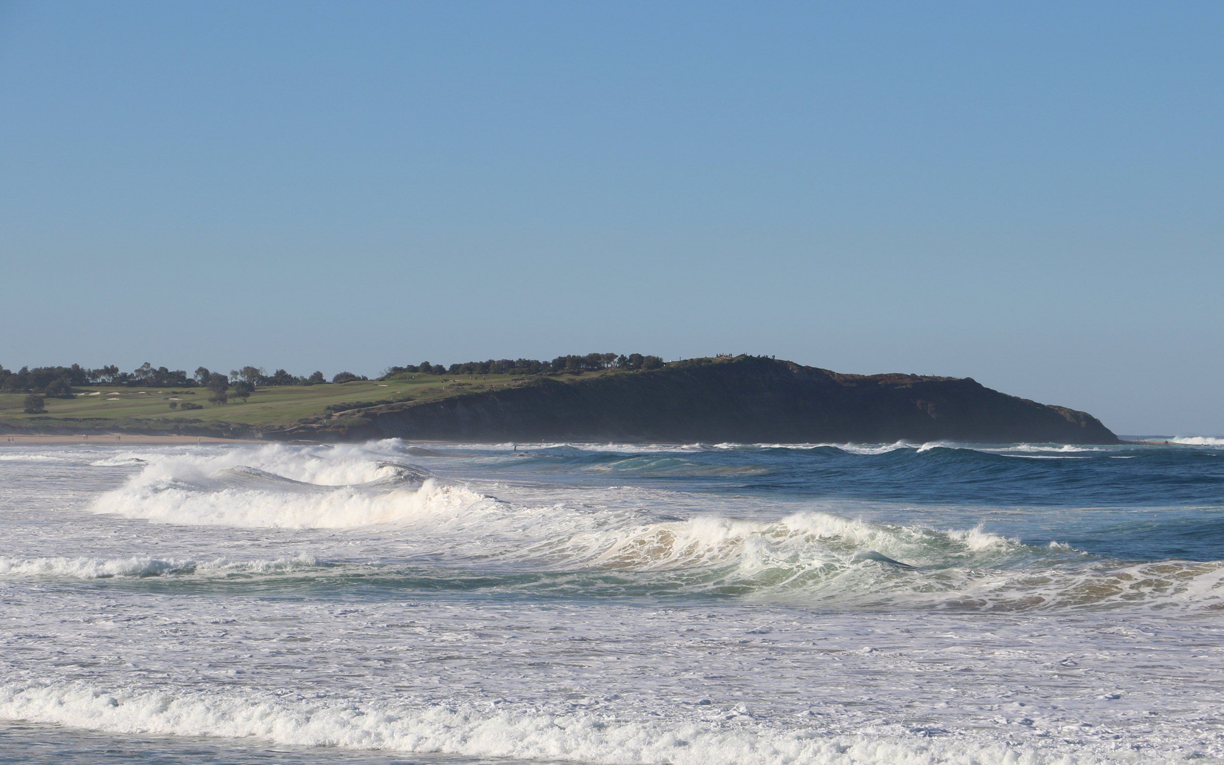 Waves breaking along Long Reef Beach, the green headland at its far end