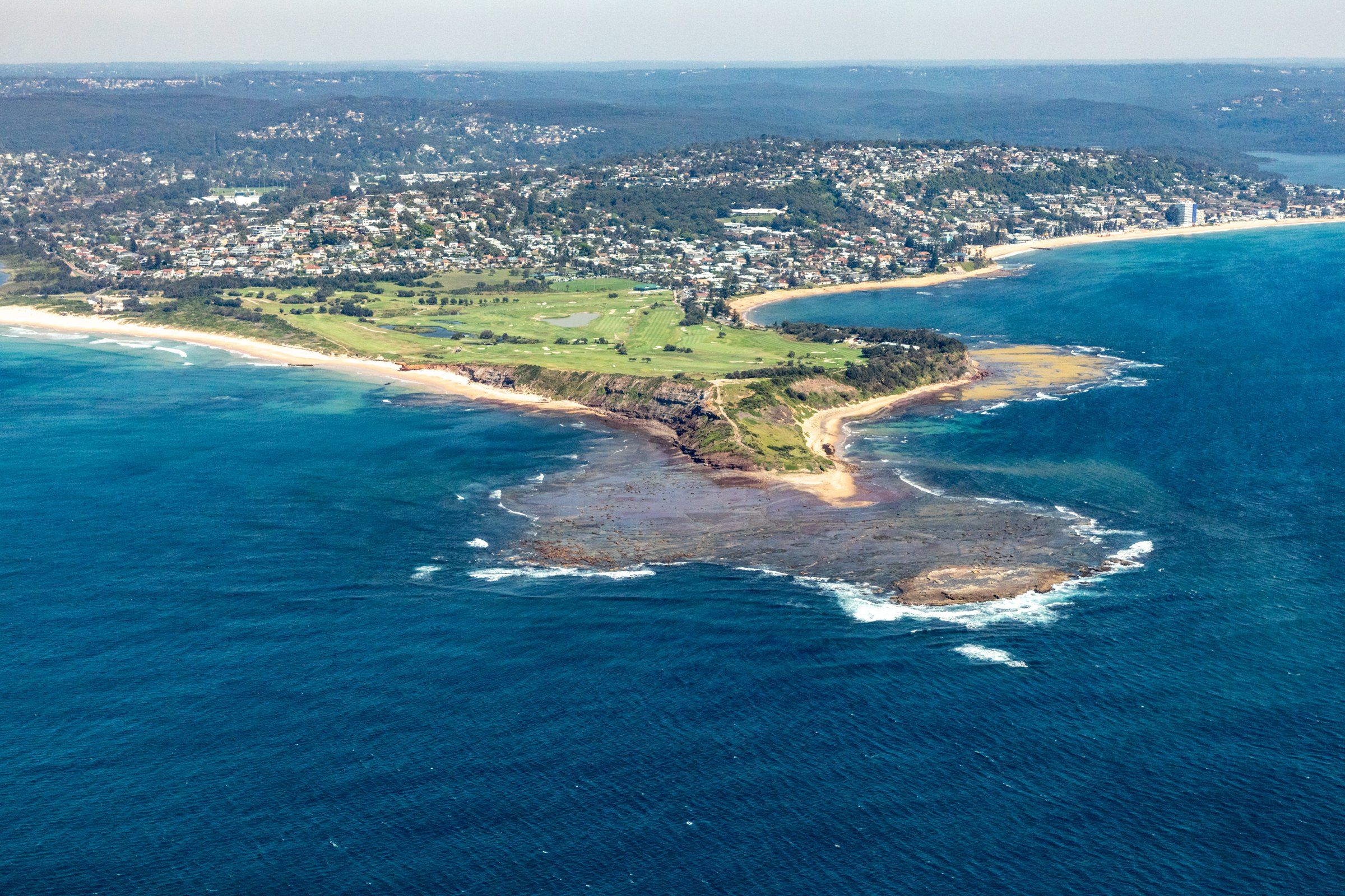 Long Reef headland from the air: the green golf course, the rock platform reaching into blue water, and the beaches either side