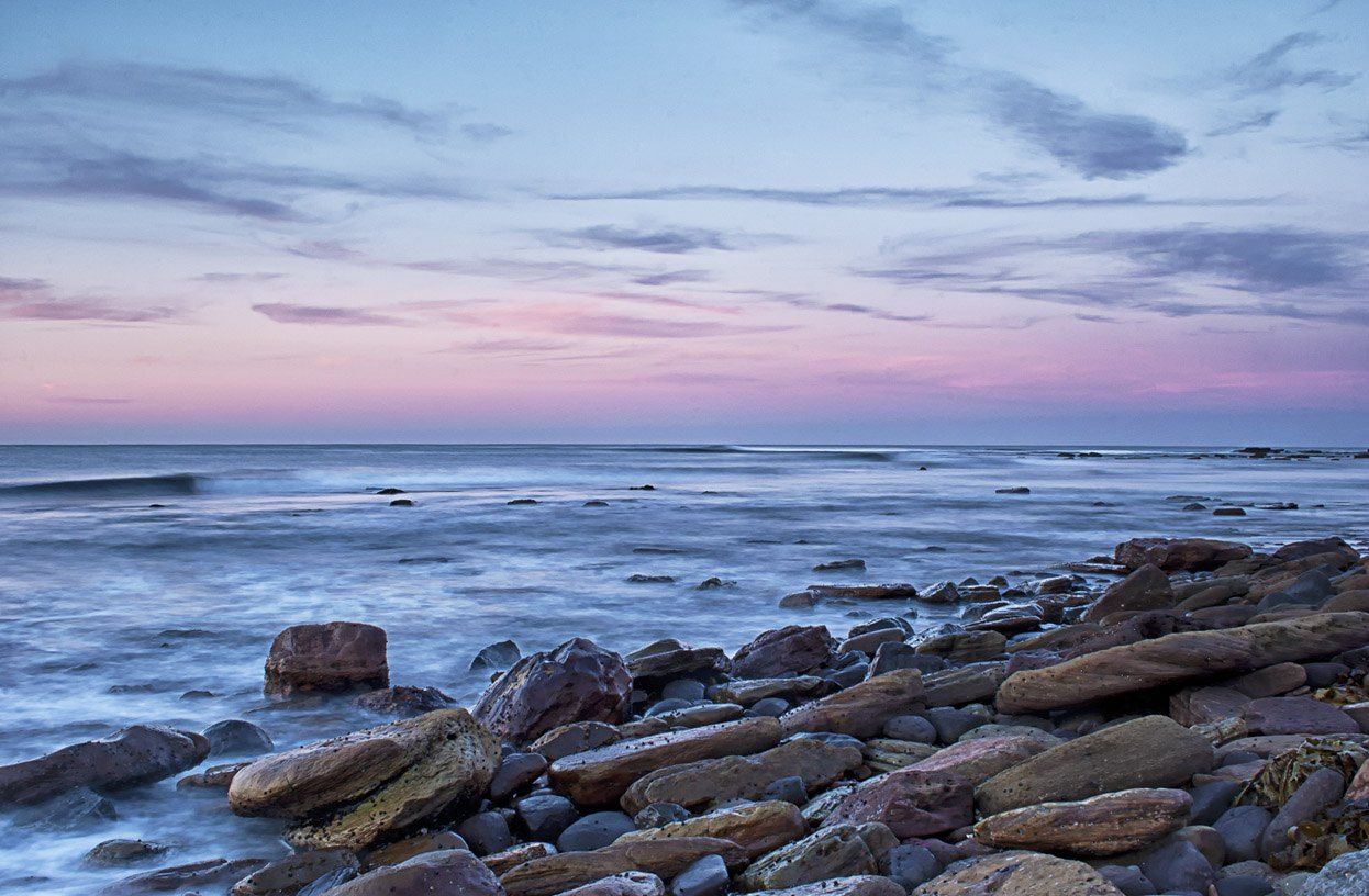 Boulders on the shore at Long Reef at dusk, the sea smoothed by a long exposure under a pink sky
