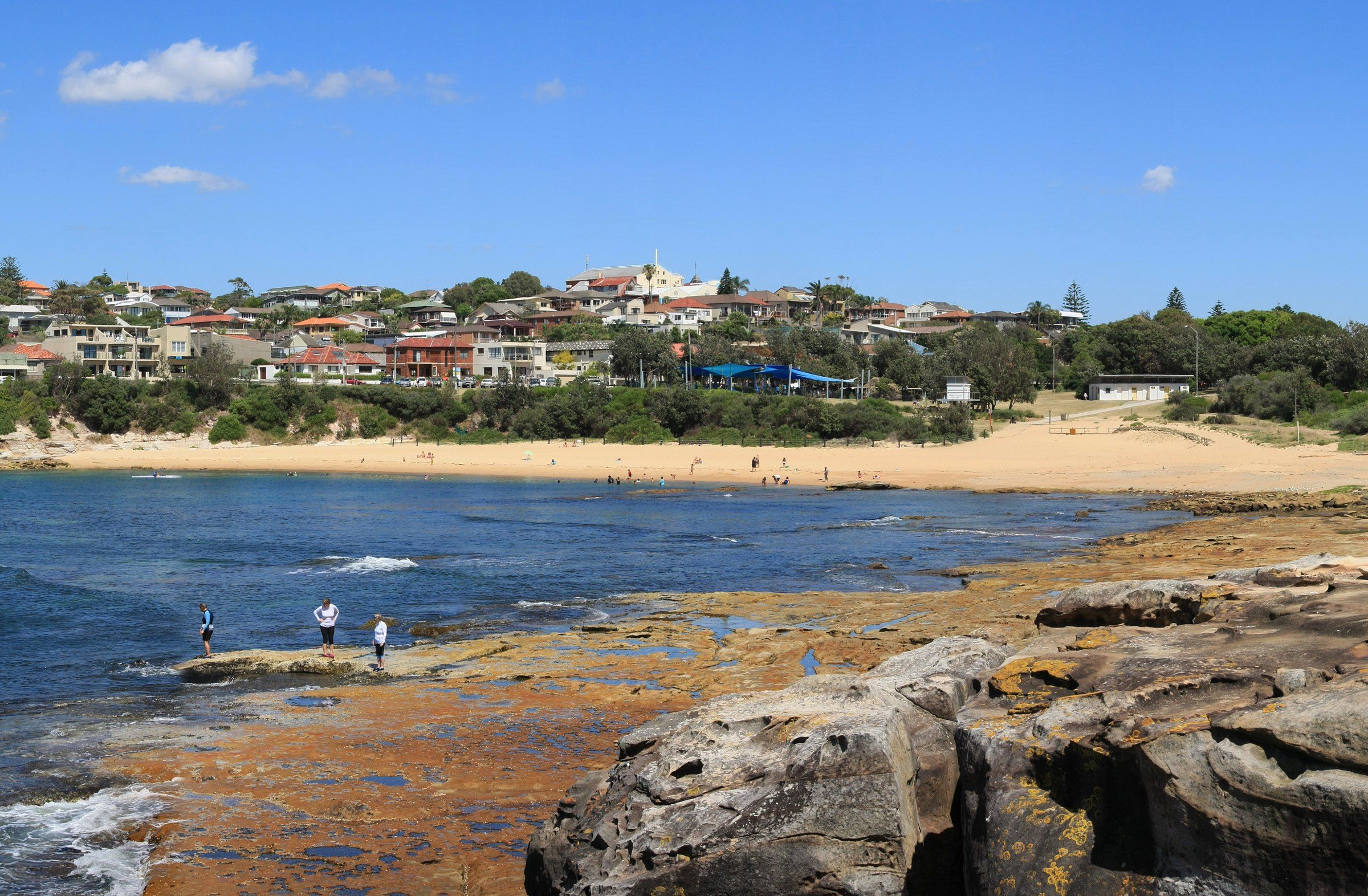 Malabar Beach across Long Bay on a sunny day, people fishing off the rocks in front and houses above the sand