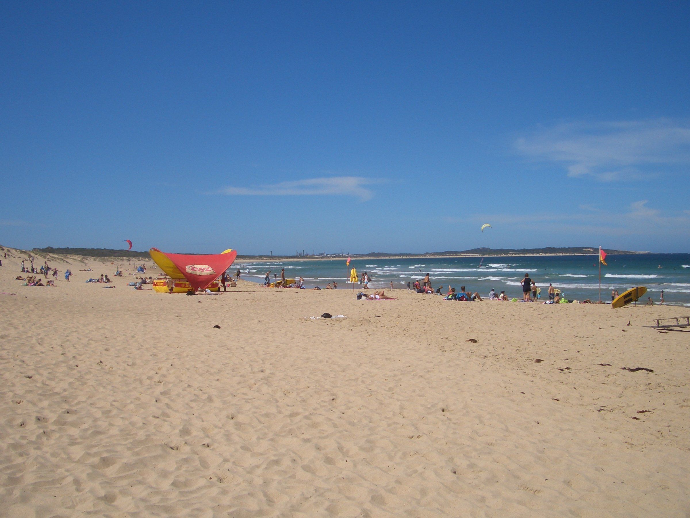 Wide, sunny sand at Wanda Beach, a lifeguard shelter and flags near the water